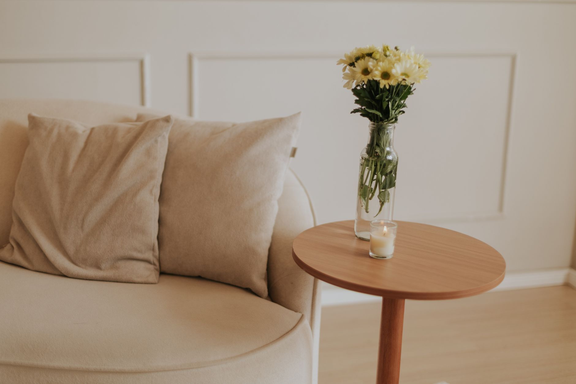 Um canto de sala de espera em tons neutros: um sofá bege, uma mesa de madeira, um vaso com flores claras e uma vela acesa.