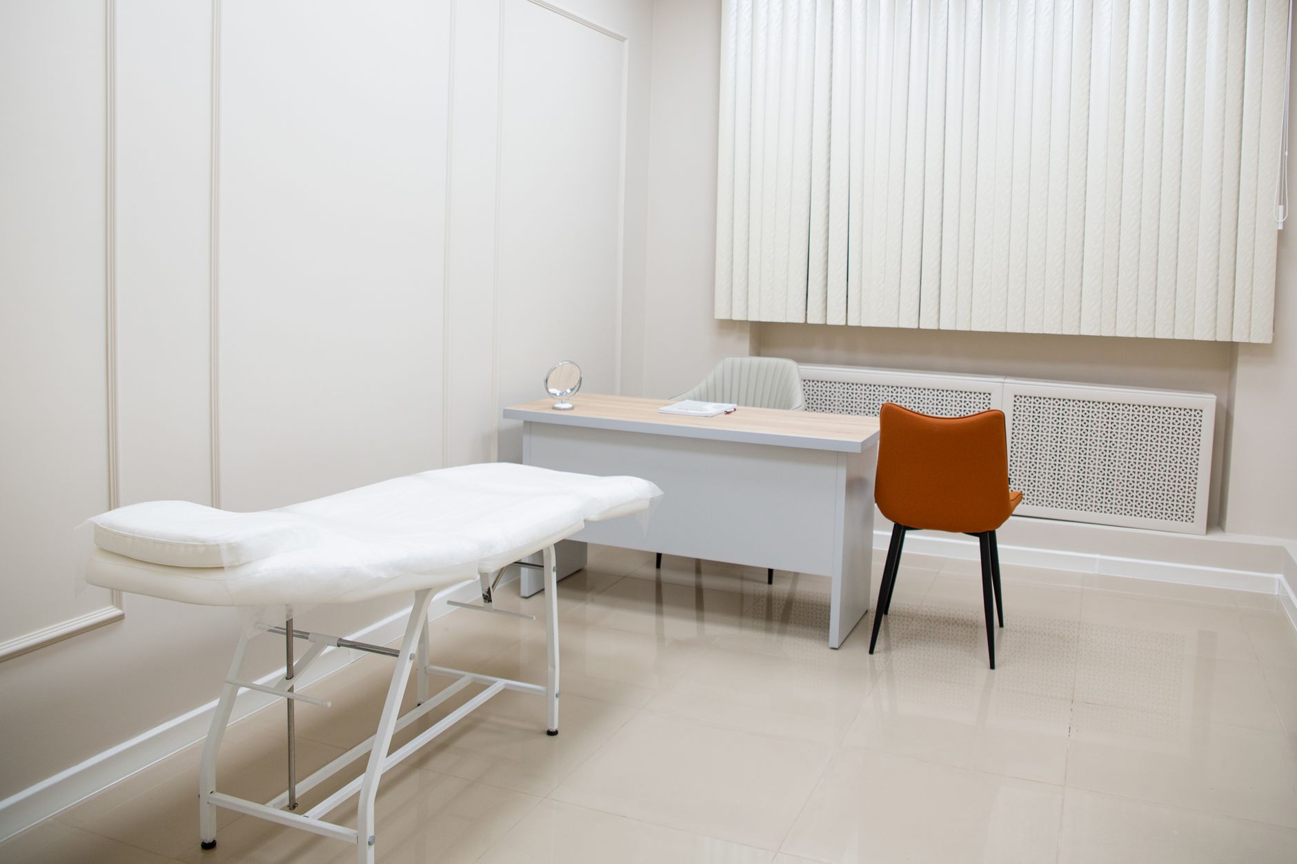 Um consultório vazio e claro, com maca forrada de branco, mesa de trabalho e uma cadeira de couro cor de terracota.